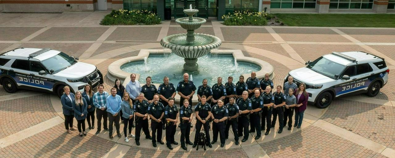 Group of police officers and some civilians posing in front of a statue for a group photo taken from above with two police cruisers on either side